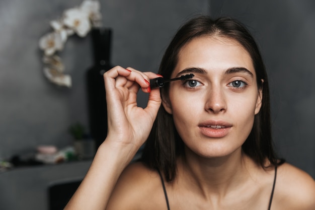 L'immagine di una giovane bella donna sveglia allegra in bagno si prende cura della sua pelle facendo il trucco con il mascara delle ciglia.