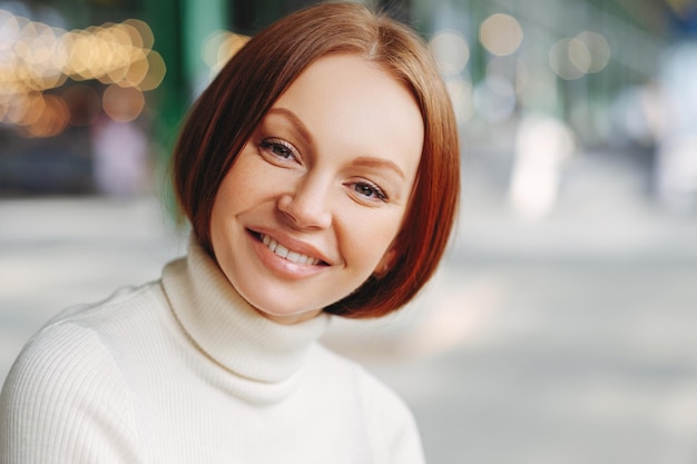 L'immagine di una giovane donna attraente con un'espressione felice guarda direttamente la fotocamera ha un aspetto soddisfatto indossa un maglione a collo alto posa su sfondo sfocato cammina fuori esprime buone emozioni