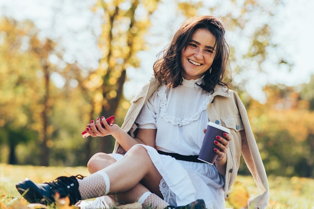 L'immagine di una giovane donna felice tiene una tazza di caffè usa e getta utilizzando lo smartphone e rilassarsi nel parco cittadino Bellissimo studente che beve caffè da asporto e naviga online sul cellulare