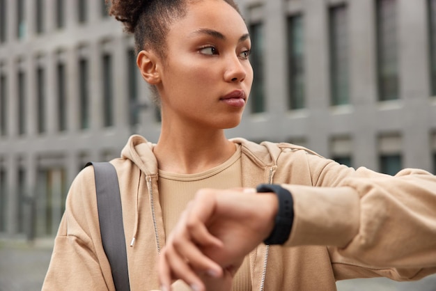 L'immagine orizzontale di una donna sportiva vestita con una felpa con cappuccio marrone indossa un orologio da polso controlla il tempo guarda seriamente le pose su uno sfondo urbano sfocato conduce uno stile di vita sano Modello femminile atletico