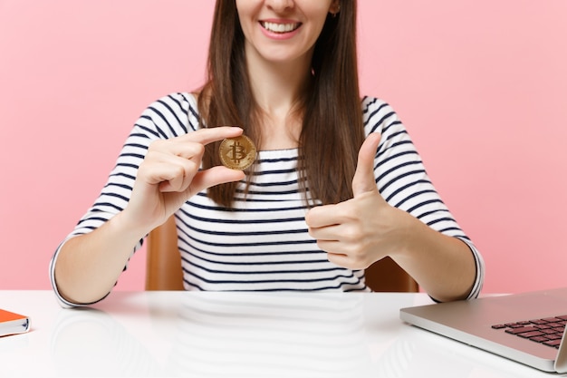 L'immagine ritagliata di una ragazza che mostra il pollice alzato tiene in mano una moneta di metallo bitcoin di colore dorato futura valuta seduta alla scrivania