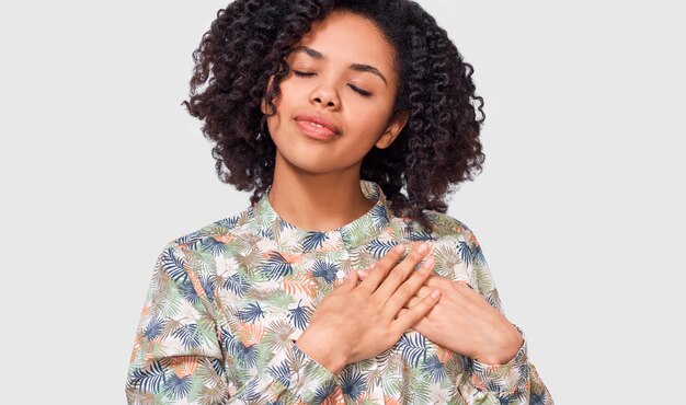 L'immagine schietta della giovane donna afroamericana tiene entrambi i palmi delle mani sul petto con gli occhi chiusi vestito con una camicia floreale isolata su sfondo bianco Emozioni della gente salute concetto di linguaggio del corpo