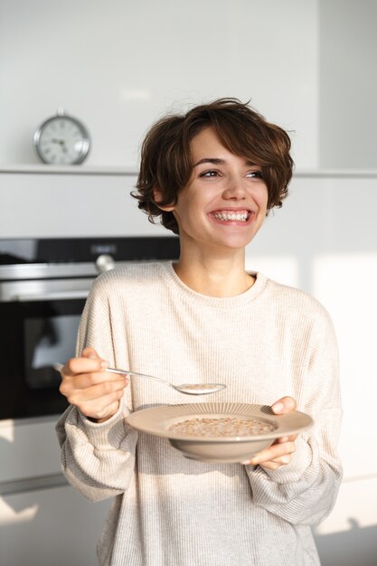 L'immagine verticale della donna castana allegra cenando e distogliendo lo sguardo mentre levandosi in piedi alla cucina