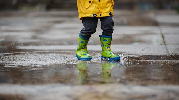 L'impermeabile giallo del bambino e gli stivali verdi cadono in una pozza d'acqua. Tempo piovoso autunnale.