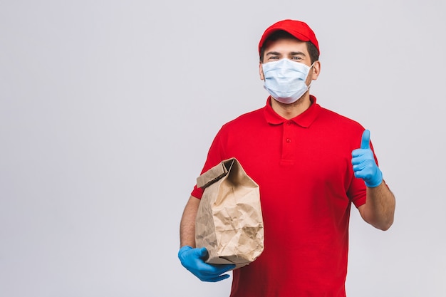 L'impiegato del fattorino in guanti della maschera di protezione dell'uniforme della maglietta dello spazio in bianco dello spiritello malevolo tiene la scatola di cartone vuota isolata sulla parete bianca. Servizio quarantena pandemia coronavirus virus 2019-ncov concept.