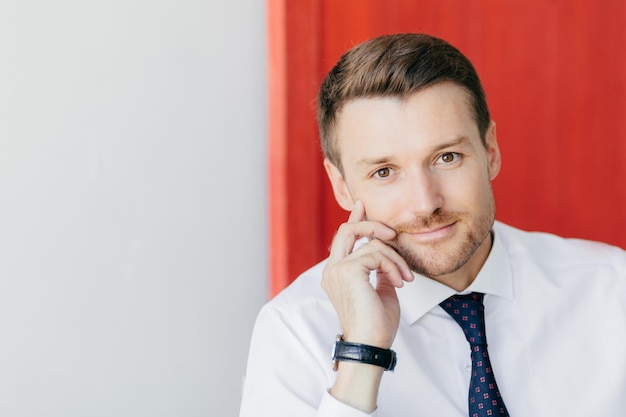 L'impiegato maschio con la barba lunga sicuro di sé in camicia bianca formale con cravatta tiene la mano sulla guancia pensa di iniziare una nuova fase nella vita pone su sfondo rosso e bianco Concetto di business