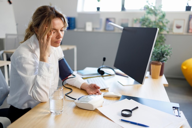 L'impiegato stanco della donna d'affari misura la pressione sanguigna mentre è seduto sul posto di lavoro