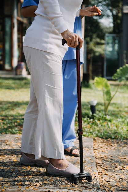 L'infermiera asiatica della giovane donna alla casa di cura si prende cura dell'uomo anziano disabile Il medico del caregiver serve la terapia fisica per il paziente anziano più anziano per esercitarsi e esercitarsi a camminare sul deambulatore o sul bastone nel cortile