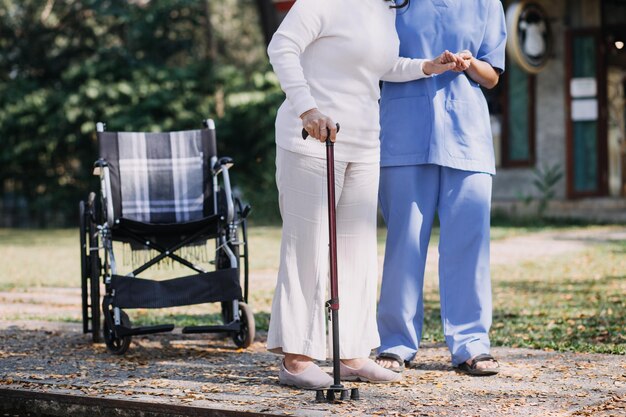 L'infermiera asiatica della giovane donna alla casa di cura si prende cura dell'uomo anziano disabile Il medico del caregiver serve la terapia fisica per il paziente anziano più anziano per esercitarsi e esercitarsi a camminare sul deambulatore o sul bastone nel cortile