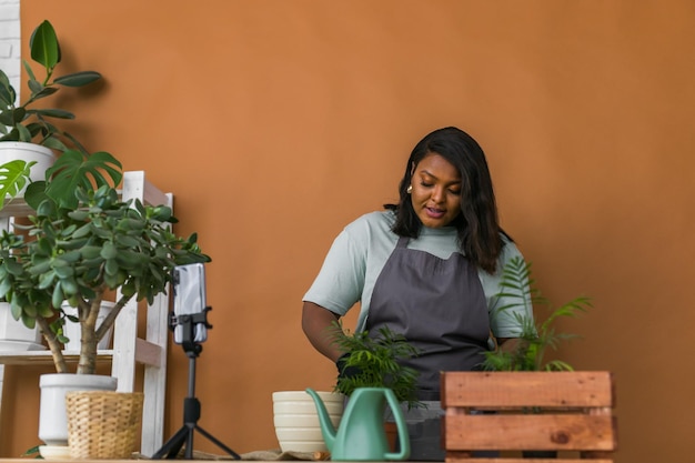 L'influencer di blogger di ragazza afroamericana lavora su riprese selfie con videocamera domestica prendersi cura delle piante domestiche e trapiantare piante in vaso di fiori Giardinaggio domestico e concetto di fiorista