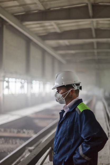 L'ingegnere minerario in casco bianco e respiratore ispeziona l'officina sporca polverosa