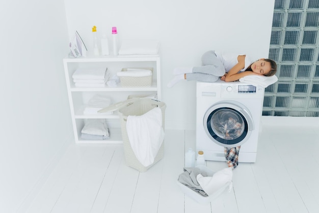 L'inquadratura di un bambino stanco che dorme sulla lavatrice usa un asciugamano morbido bianco poiché il cuscino ha sogni piacevoli il cestino e il bacino sul pavimento bianco si sente affaticato dopo aver caricato i vestiti nella lavatrice essendo a casa