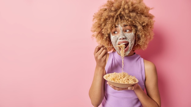 L'inquadratura interna di una bella donna dai capelli ricci applica una maschera di bellezza per ridurre i punti neri e le linee sottili mangia pasta appetitosa distoglie lo sguardo si erge contro lo spazio vuoto della parete rosa dello studio per il tuo promo