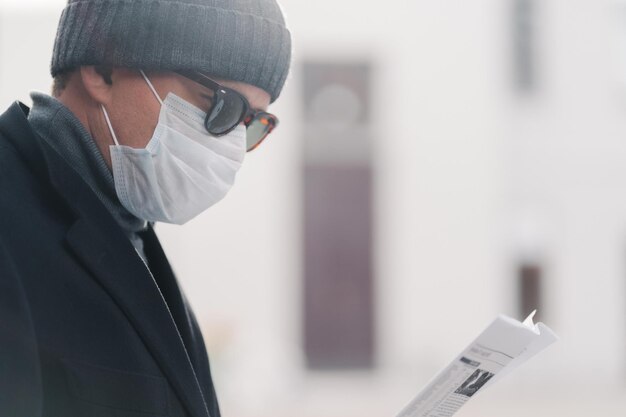 L'inquadratura laterale dell'uomo con occhiali da sole e maschera protettiva legge il giornale pose all'aperto in strada scopre notizie dalla stampa durante l'epidemia di coronavirus e il virus respiratorio in quarantena