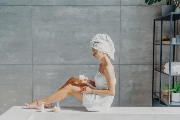 L'inquadratura orizzontale di una giovane donna europea rilassata applica una crema idratante per il corpo sulle gambe avvolte in un asciugamano da bagno ha un sorriso tenero, una pelle sana e rinfrescata dopo aver preso le pose del bagno in un bagno accogliente