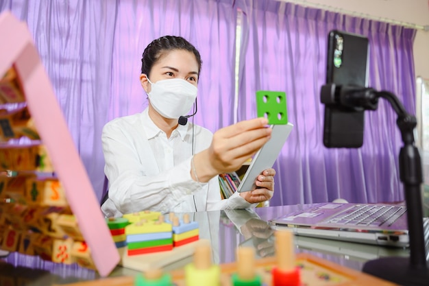 L'insegnante asiatica che indossa una maschera insegna agli studenti a studiare online attraverso lo schermo di un computer utilizzando un sistema di videoconferenza online per l'istruzione