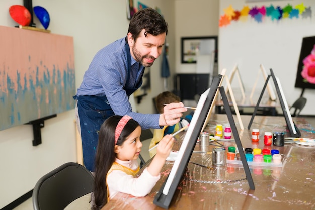 L'insegnante maschio sorridente è felice di aiutare una bambina a dipingere su una tela durante una lezione d'arte per bambini