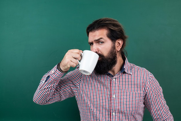 L'insegnante maturo dell'uomo barbuto beve il caffè nell'aula della scuola con la lavagna, mattina