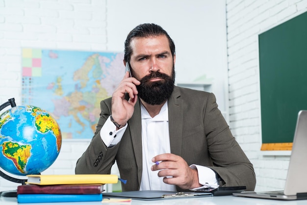 L'insegnante maturo serio parla al telefono l'uomo barbuto brutale lavora in classe e lo smartphone hols si prepara per l'esame online docente universitario durante la lezione torna a scuola comunicazione di educazione formale