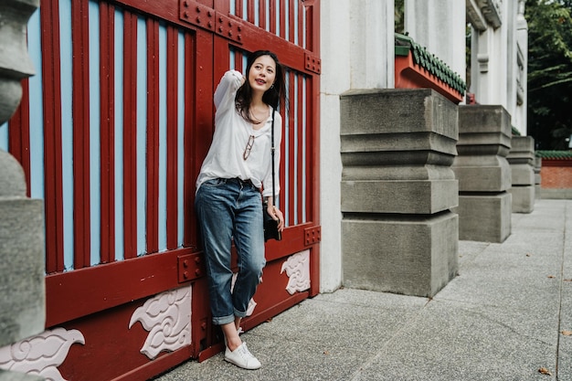 l'intera lunghezza della giovane turista asiatica coreana con la borsa si muove i capelli e sta in piedi contro la porta rossa dal vecchio edificio cinese. il viaggiatore felice della ragazza si rilassa all'aperto aspettando qualcuno all'ingresso.