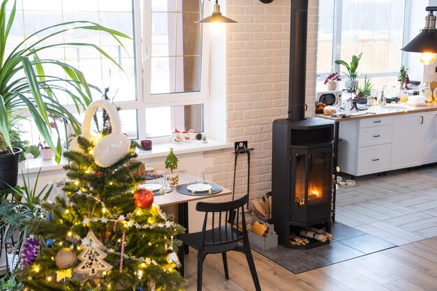 L'interno festivo della casa è decorato per Natale e Capodanno in stile loft con stufa nera camino albero di Natale Calda sala studio con cucina bianca che brucia legna accogliente e riscaldamento di casa