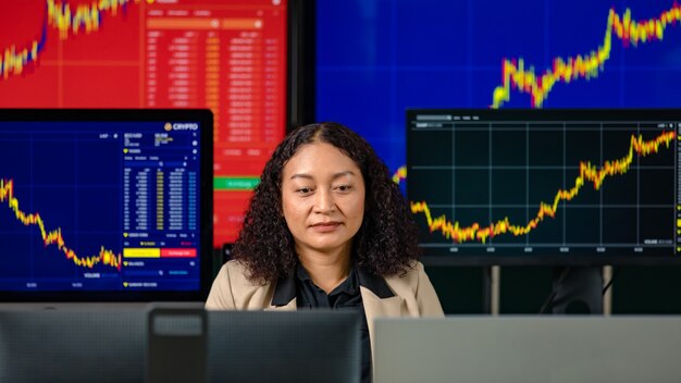 L'investitore professionista di successo femminile si siede sorridendo guarda la telecamera circondata dallo schermo del monitor del computer con il grafico grafico e il rapporto di analisi della criptovaluta bitcoin nella sala di negoziazione.