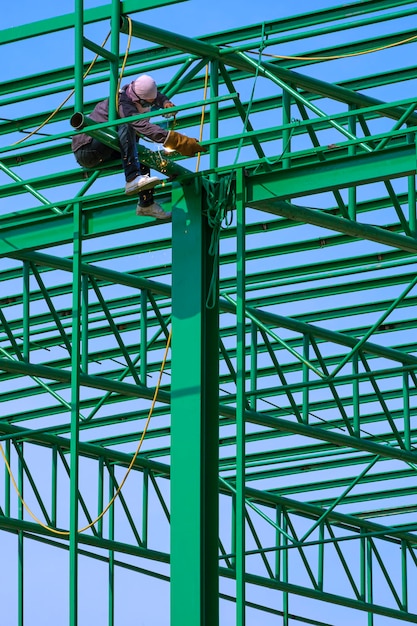 L'operaio edile asiatico sta saldando il metallo sulla struttura del magazzino del tetto verde contro il cielo blu