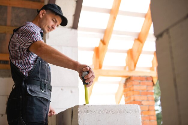 L'operaio edile in cantiere misura la lunghezza dell'apertura della finestra e il muro di mattoni con metro a nastro Cottage sono fatti di blocchi di cemento poroso abiti da lavoro tuta e berretto da baseball