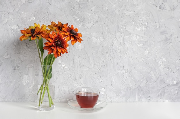 L'ora del tè. Tè nero in tazza trasparente con piattino e bouquet di fiori d'arancio coneflowers in vaso sul tavolo