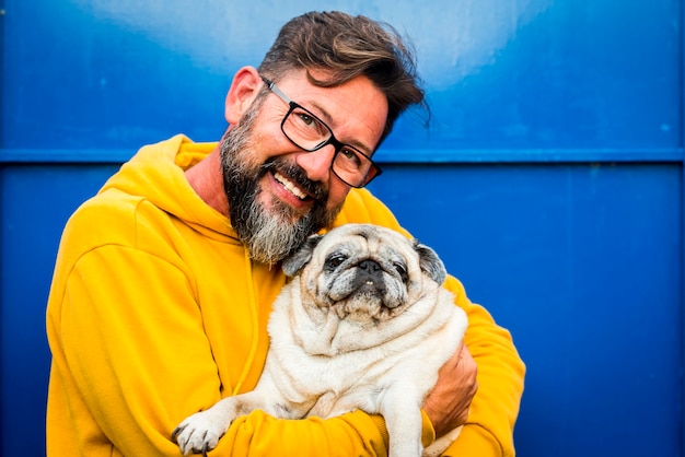 L'uomo adulto allegro sorride e abbraccia con amore il suo vecchio cane carlino in un ritratto con colore giallo e blu - le persone con animali e occhiali si godono il concetto di pet therapy