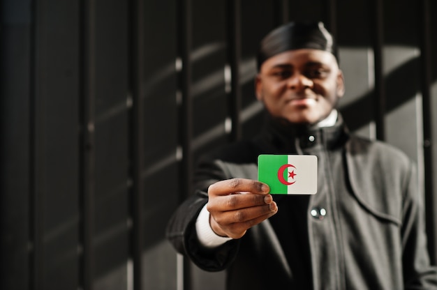 L'uomo africano indossa durag nero tenere la bandiera dell'Algeria a portata di mano isolato muro scuro.