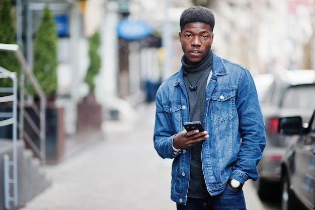 L'uomo africano indossa la giacca di jeans posata all'aperto in piedi contro l'auto nera con il telefono cellulare a portata di mano