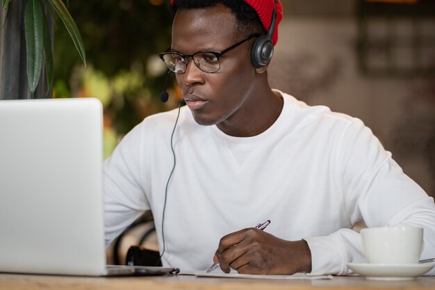 L'uomo afro indossa le cuffie, lavora online in remoto al bar, prende appunti, guarda il webinar.