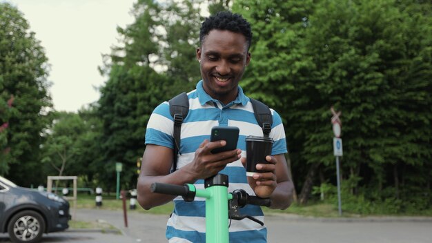 L'uomo afro usa il supporto per smartphone con scooter elettrico e sorseggia una bevanda calda per andare l'uomo usa un'app sul suo telefono per noleggiare uno scooter elettrico trasporto ecologico urbano all'aperto