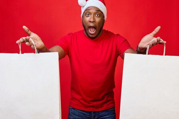 L'uomo afroamericano con la bocca aperta per la sorpresa in un cappello da Babbo Natale tiene in mano dei sacchetti di carta