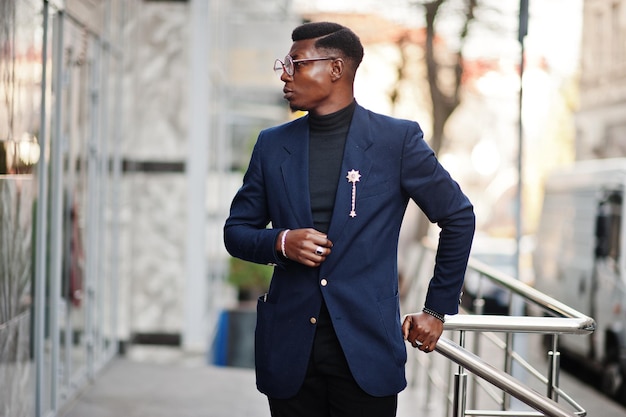 L'uomo afroamericano dall'aspetto sorprendente indossa un blazer blu con spilla a collo alto nero e occhiali posati in strada Ragazzo nero alla moda