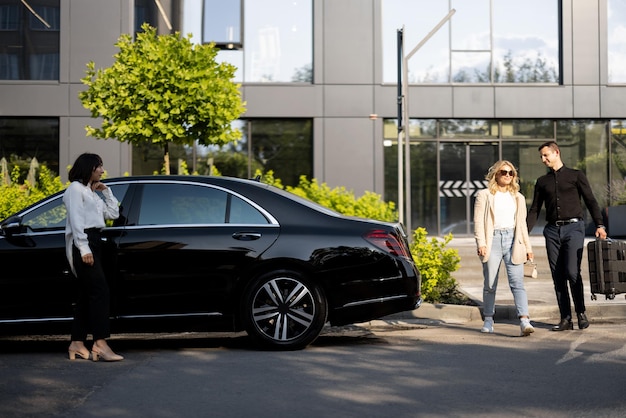 L'uomo aiuta una donna d'affari a portare la valigia in un'auto di lusso da un edificio per uffici o un hotel L'autista femminile attende vicino a un'auto Concetto di viaggi d'affari e servizi di trasferimento di lusso