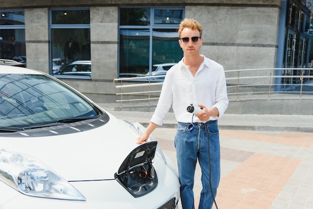 L'uomo alla moda con la tazza di caffè in mano inserisce la spina nella presa di ricarica dell'auto elettrica