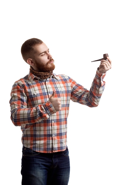 L'uomo alla moda della barba rossa in camicia a quadri con il pollice in su mostra la pipa fumante su sfondo bianco