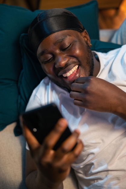 L'uomo allegro giace sul divano a fissare il telefono tocca mano a faccia ridendo mostrando denti bianchi