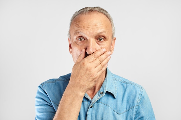 L'uomo anziano dai capelli grigi scioccato e sorpreso chiude spaventosamente la bocca con la mano isolata su sfondo bianco, concetto di emozioni umane