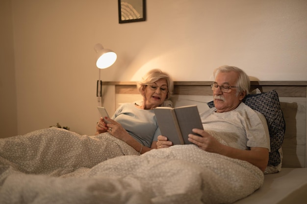 L'uomo anziano e sua moglie si rilassano a letto la sera L'uomo sta leggendo un libro mentre la donna usa il cellulare