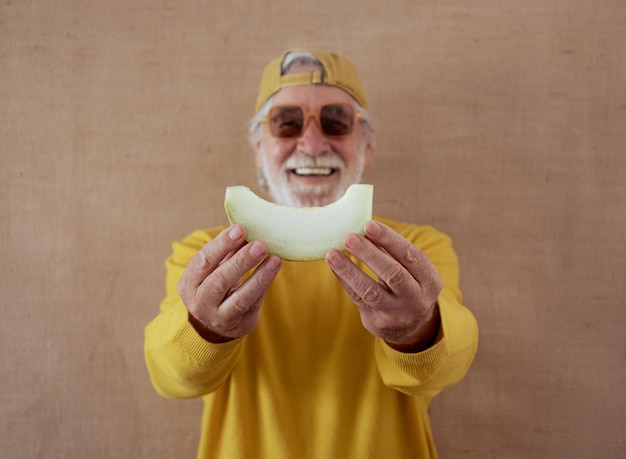 L'uomo anziano sfocato in camicia gialla e cappello isolato su uno sfondo chiaro tiene in mano una fetta di melone pronta per essere mangiata