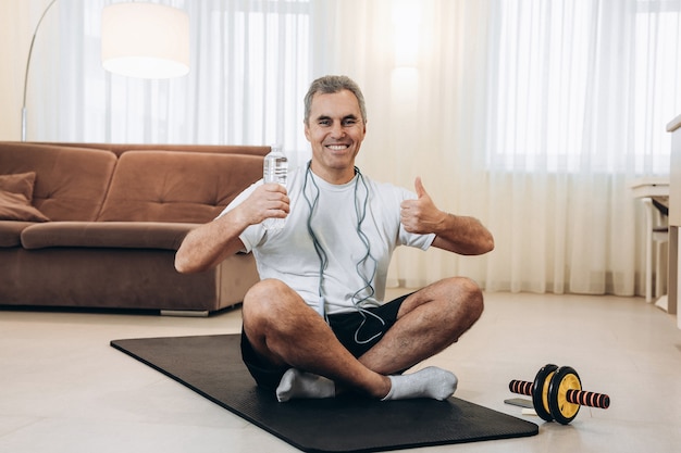 L'uomo anziano tiene in mano una bottiglia d'acqua, sorride con i denti, si siede sulla mappa yoga nera e pollice in alto