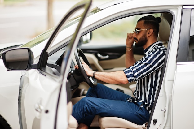 L'uomo arabo di successo indossa una camicia a righe e occhiali da sole posa al volante della sua suv bianca. Uomini arabi alla moda in trasporto.