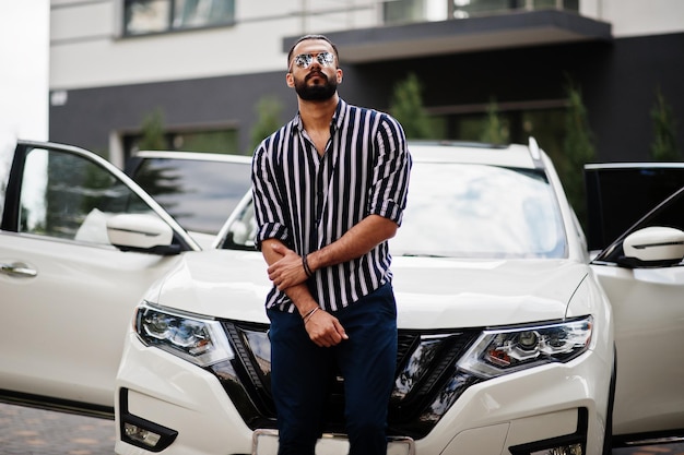 L'uomo arabo di successo indossa una camicia a righe e occhiali da sole posa vicino alla sua suv bianca. Uomini arabi alla moda in trasporto.