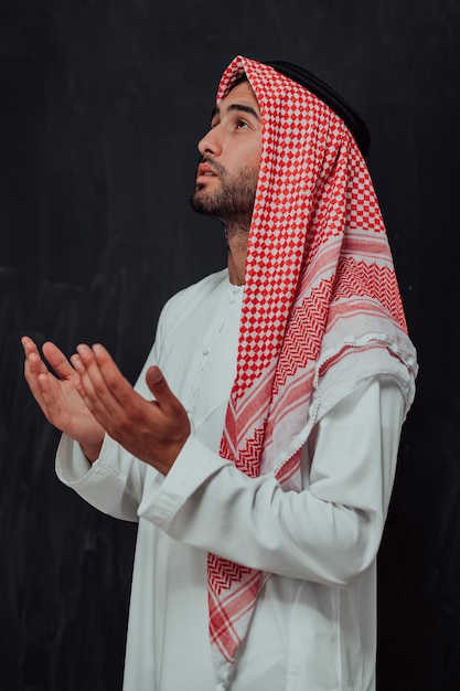L'uomo arabo in abiti tradizionali che fa la preghiera tradizionale a Dio, tiene le mani nel gesto di preghiera davanti alla lavagna nera che rappresenta la moda moderna dell'islam e il concetto di ramadan kareem.