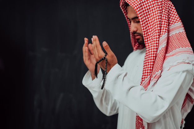 L'uomo arabo in abiti tradizionali che fa la preghiera tradizionale a Dio, tiene le mani nel gesto di preghiera davanti alla lavagna nera che rappresenta la moda moderna dell'islam e il concetto di ramadan kareem.