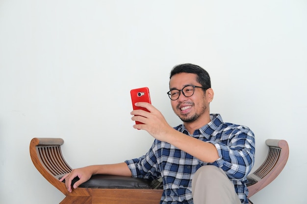 L'uomo asiatico adulto seduto si rilassa mentre guarda al suo handphone con un'espressione felice