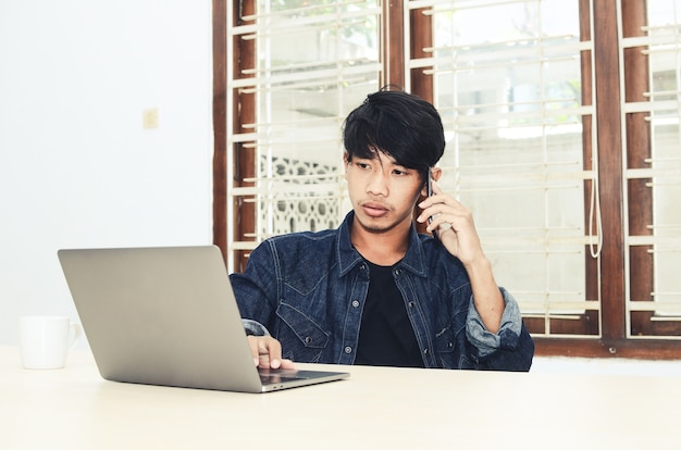 L'uomo asiatico con la camicia nera seduto davanti al laptop sta parlando al telefono